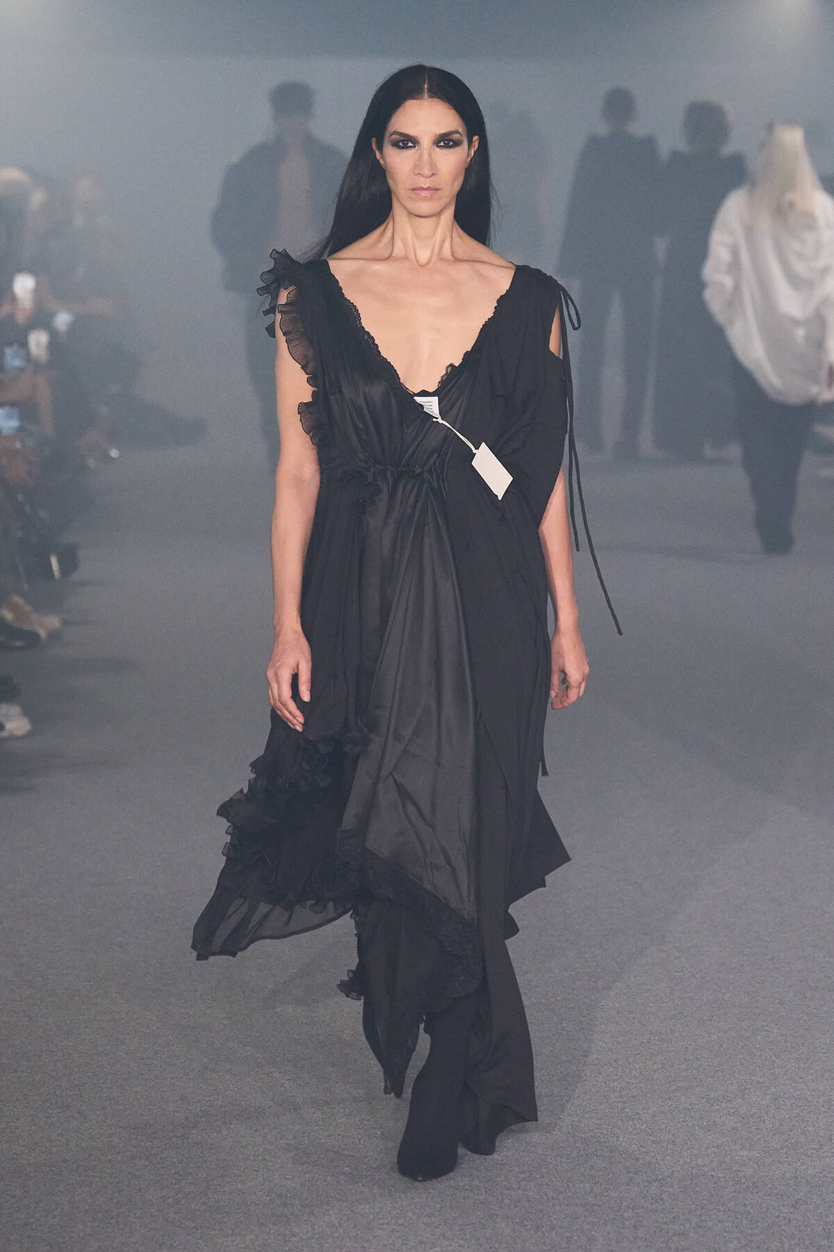 Model walking the runway in a black, flowing gown with ruffled lace details and a white tag on the dress.