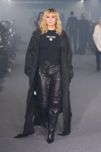 Model walking a smoky fashion show wearing an all-black outfit: long coat, POLIZEI logo tee, and black leather pants with heeled boots.