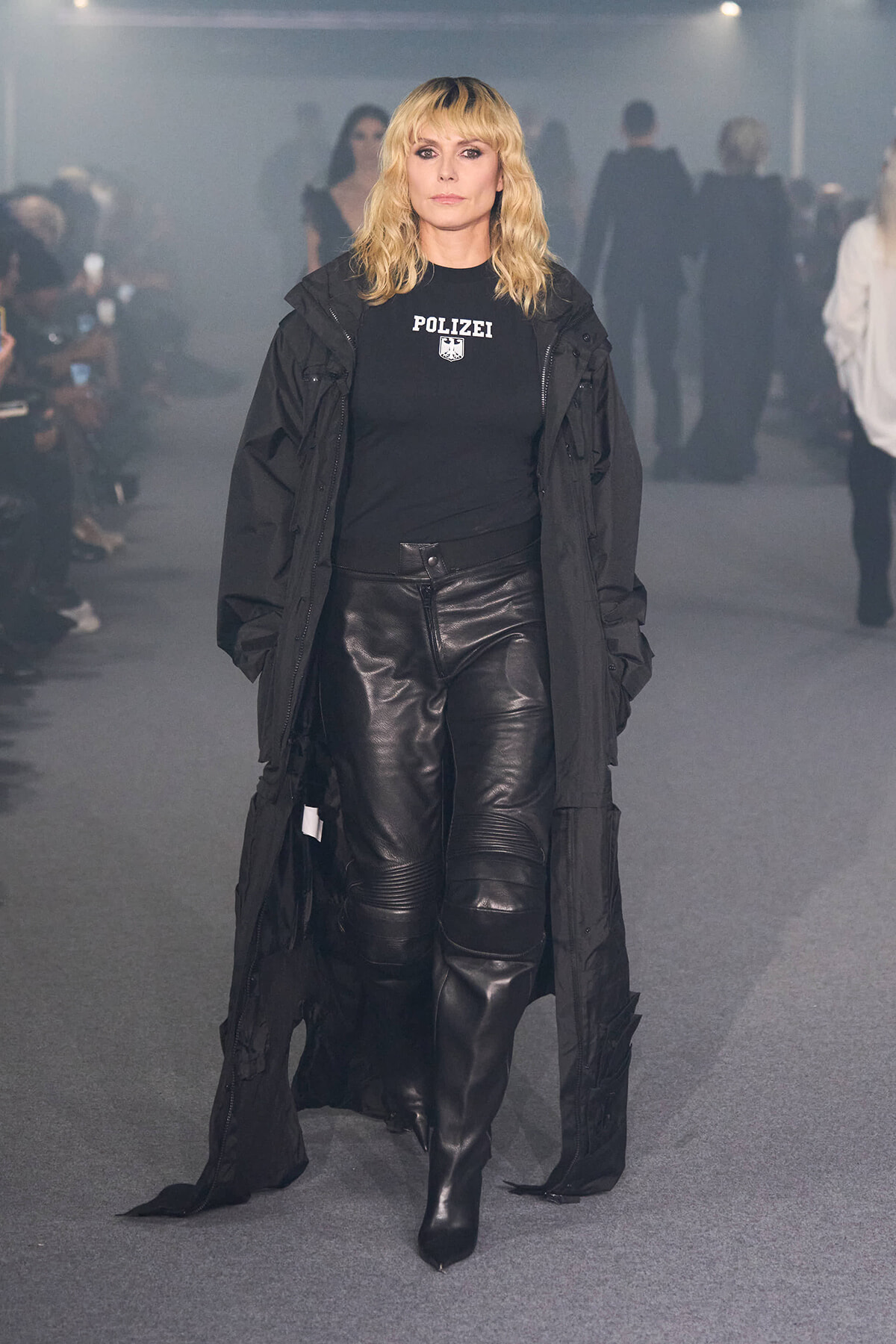 Model walking a smoky fashion show wearing an all-black outfit: long coat, POLIZEI logo tee, and black leather pants with heeled boots.