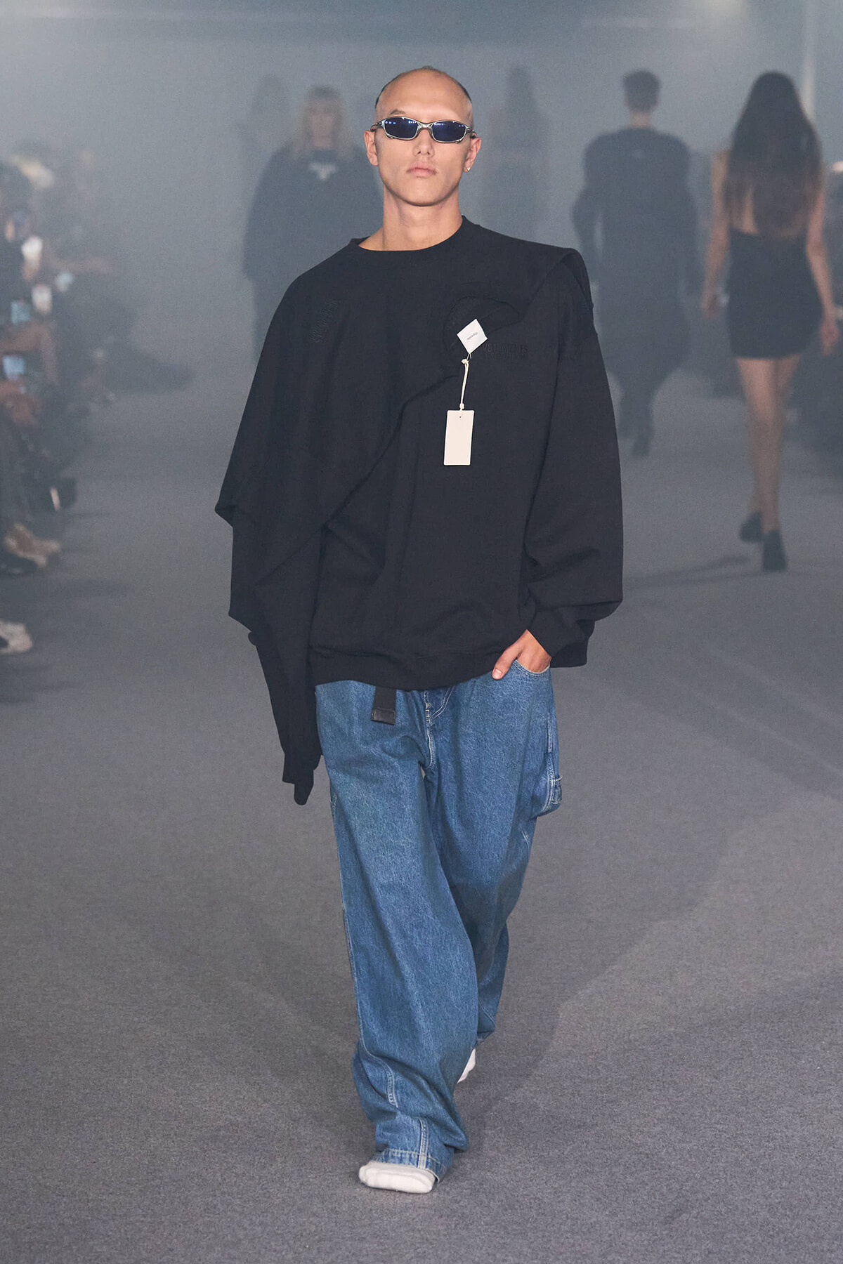 Male model on a runway wearing an oversized black top, baggy blue jeans, white sneakers, and dark sunglasses with a tag hanging from the chest.