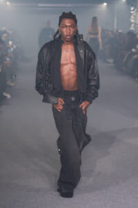 Male model walking the runway wearing a black leather jacket, open to reveal a bare chest and jeans; confident pose with hands in pockets in a dim, smoky fashion show setting.
