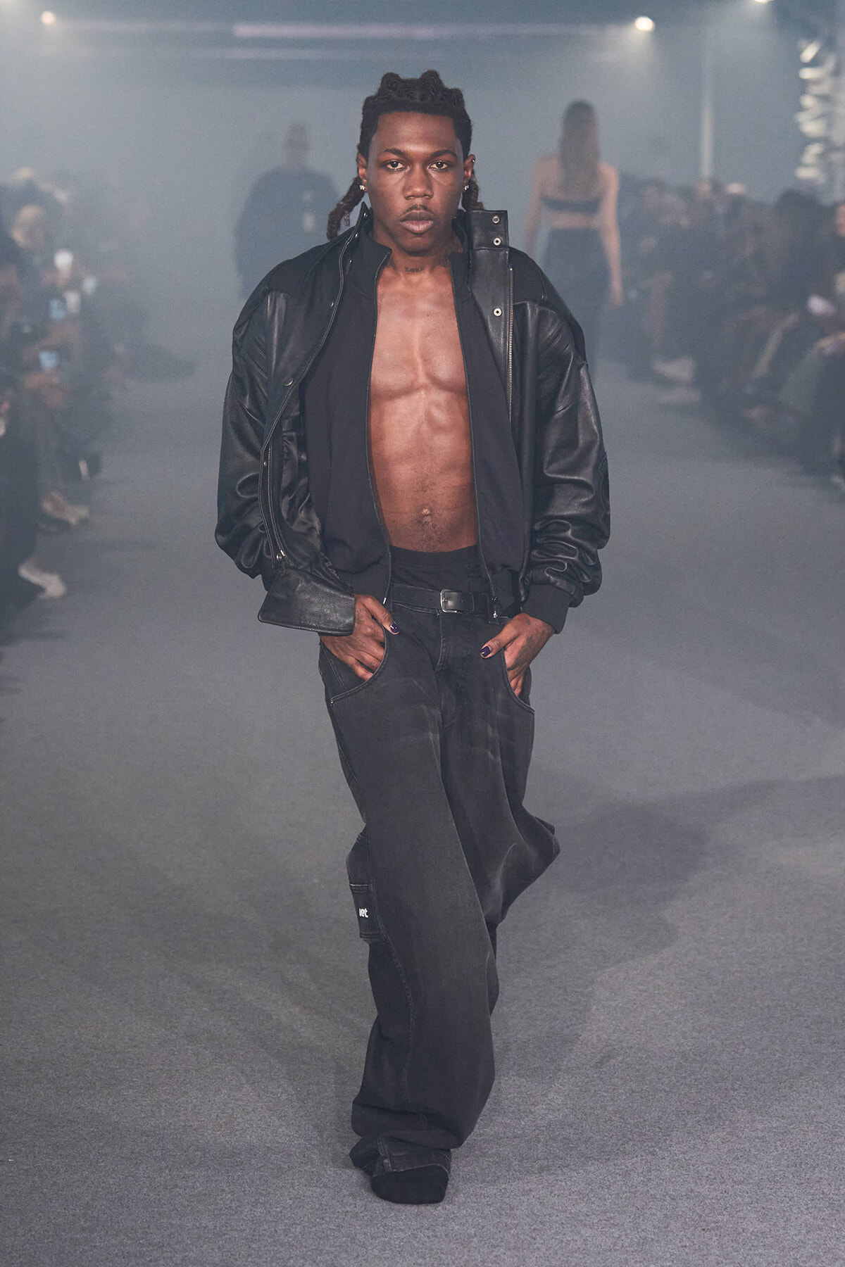 Male model walking the runway wearing a black leather jacket, open to reveal a bare chest and jeans; confident pose with hands in pockets in a dim, smoky fashion show setting.