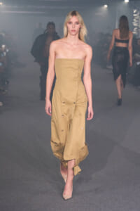 Model walking runway in a strapless, tan midi dress with large button accents and ruched gathered hem, nude heels.
