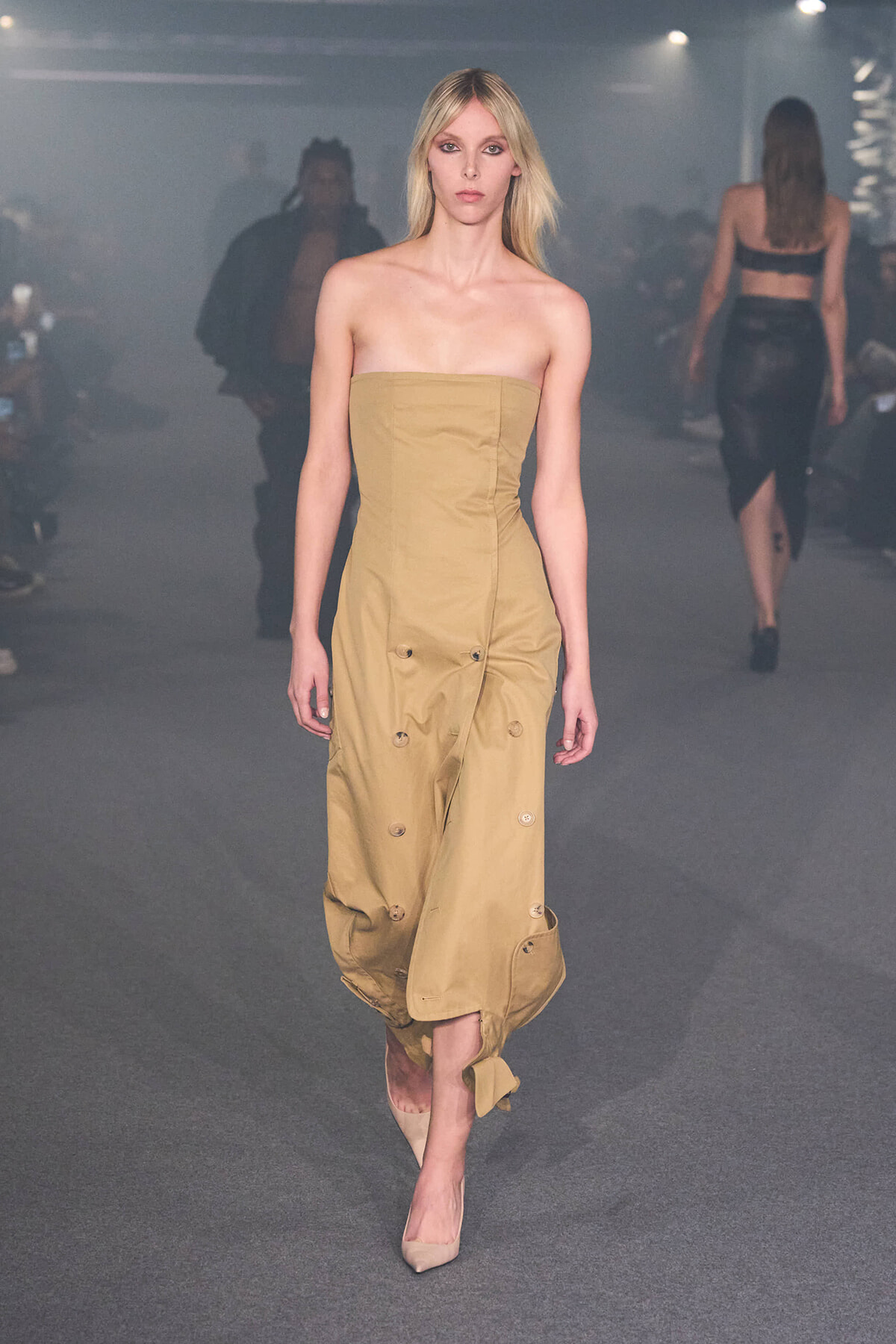 Model walking runway in a strapless, tan midi dress with large button accents and ruched gathered hem, nude heels.