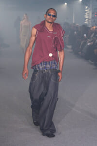 Male model on a fashion runway wearing a maroon sleeveless top, blue plaid waistband, and oversized denim jeans with sunglasses in a smoky venue.