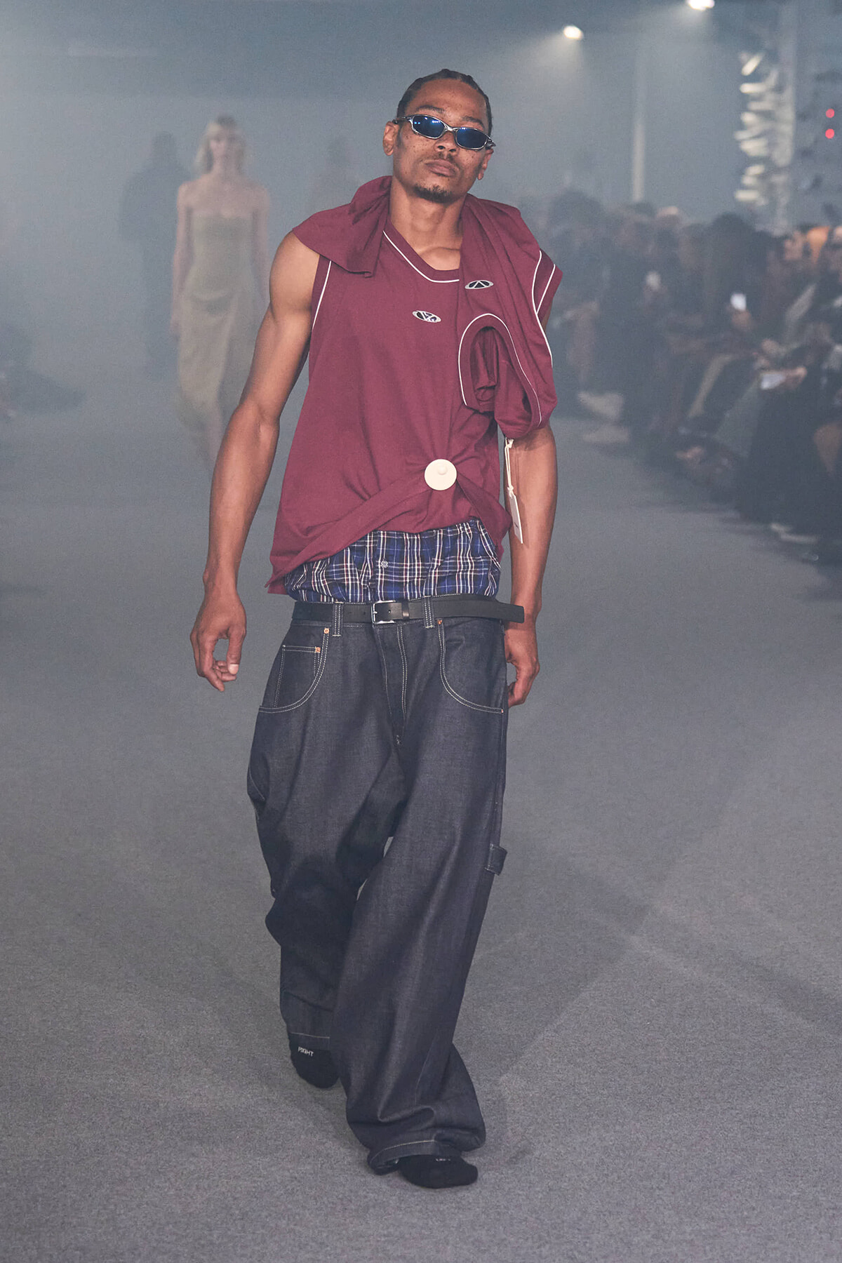 Male model on a fashion runway wearing a maroon sleeveless top, blue plaid waistband, and oversized denim jeans with sunglasses in a smoky venue.