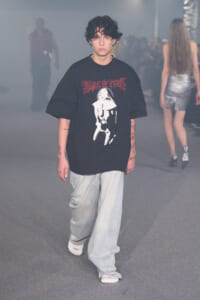 Young man with curly hair walks the runway in an oversized black graphic tee and light jeans amid a smoky backstage setting.