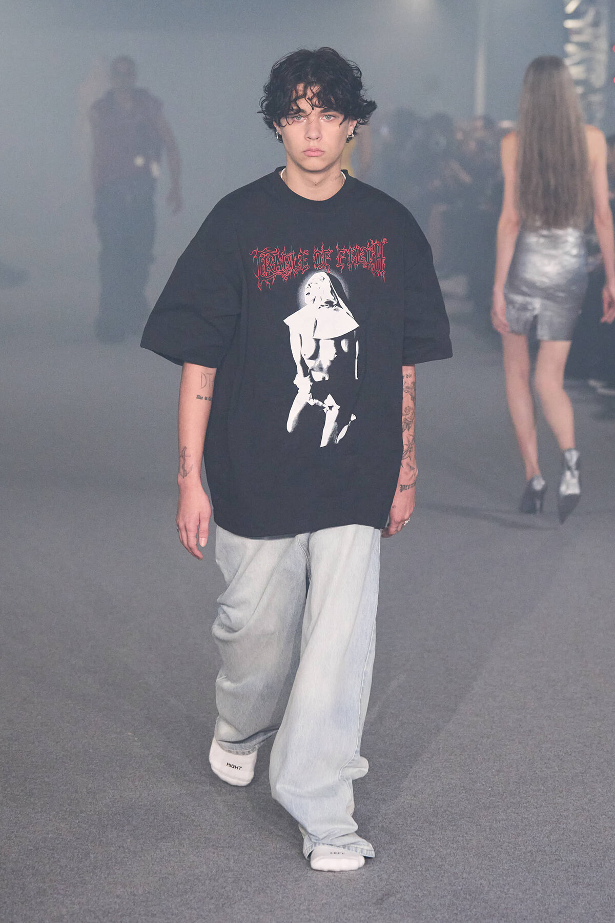 Young man with curly hair walks the runway in an oversized black graphic tee and light jeans amid a smoky backstage setting.