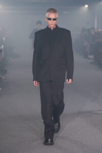 Male model walking a runway in a black suit and sunglasses amid smoky lighting on a fashion show catwalk.