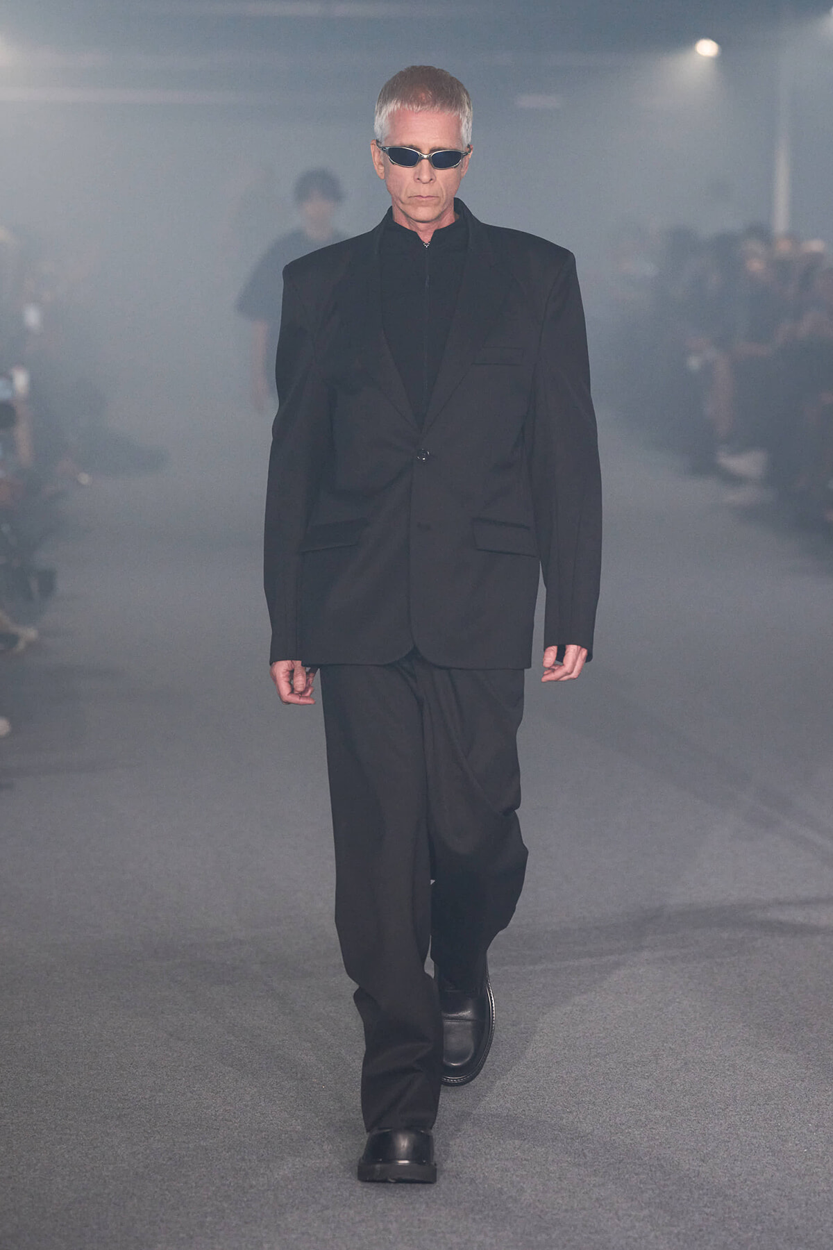 Male model walking a runway in a black suit and sunglasses amid smoky lighting on a fashion show catwalk.