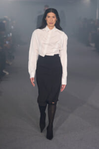 Model walking a fashion show runway in a white button-down shirt tucked into a black midi skirt with black ankle boots.