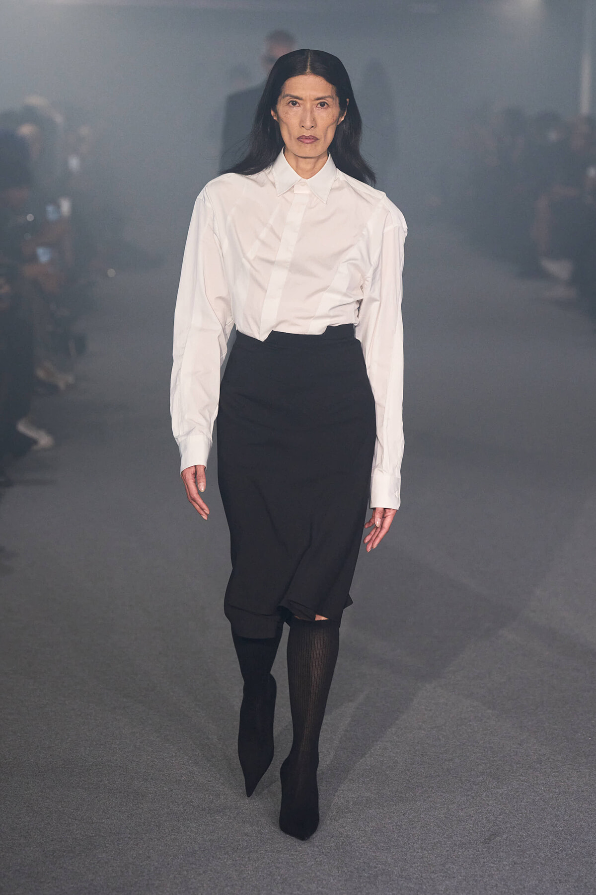 Model walking a fashion show runway in a white button-down shirt tucked into a black midi skirt with black ankle boots.