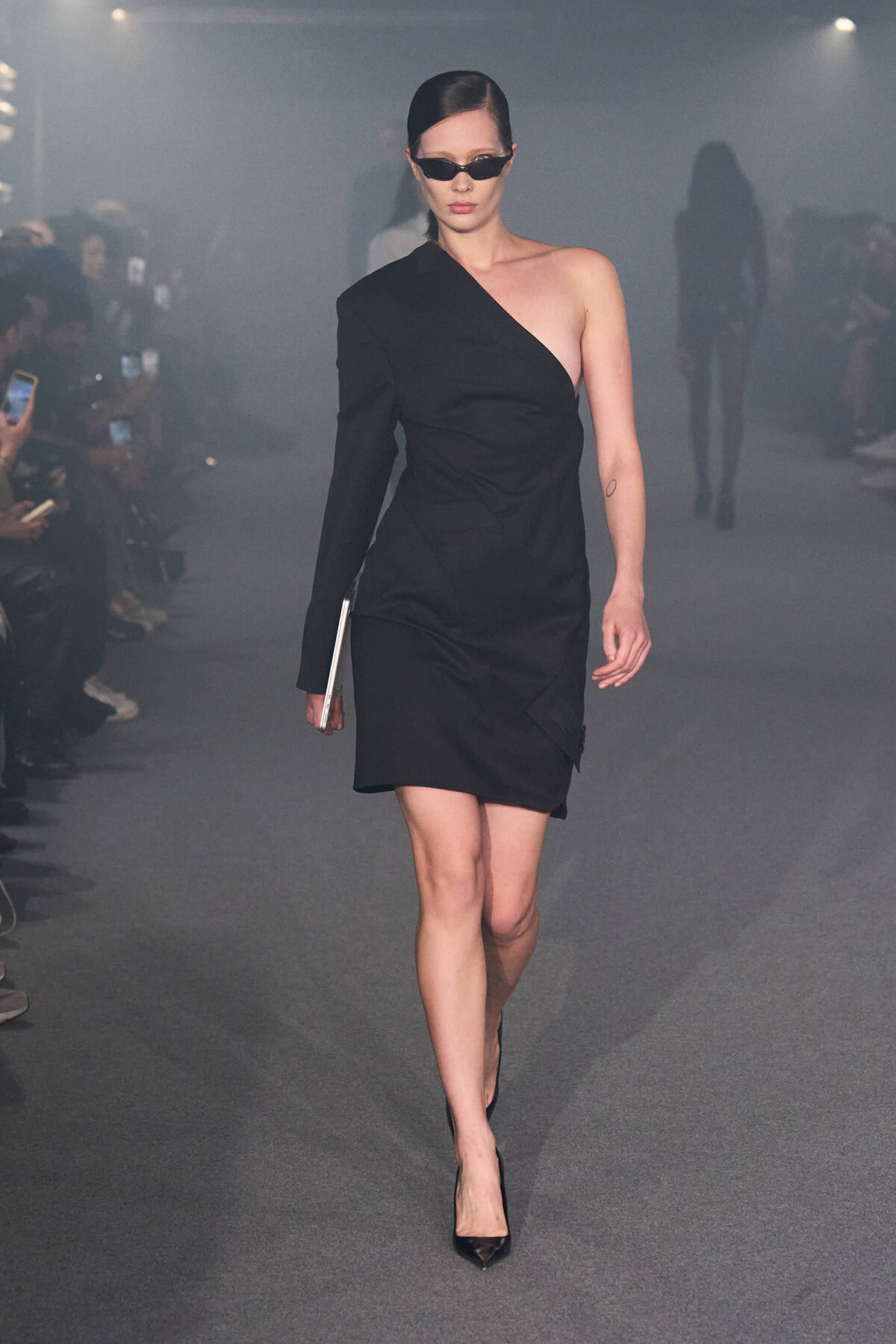 Runway model in a black one-shoulder dress with sunglasses, carrying a small clutch and wearing pointed heels.
