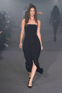 Model walking runway in a strapless black dress with a front slit, gray backstage mist in the background.