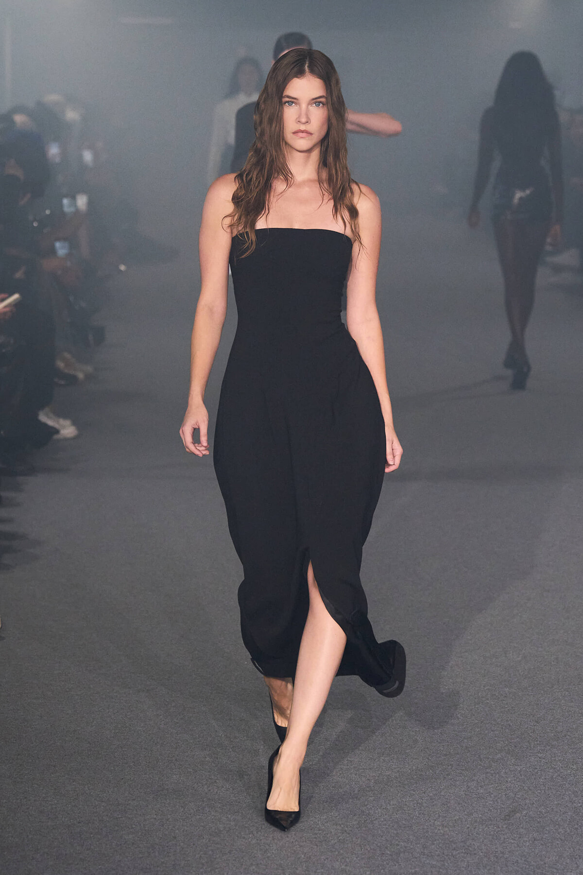 Model walking runway in a strapless black dress with a front slit, gray backstage mist in the background.