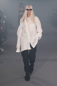 Model with long white hair wearing an oversized white shirt and black pants on a smoky runway, sunglasses on.