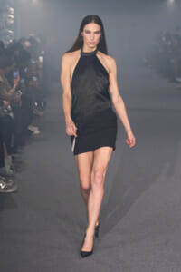 Model walking a fashion runway in a black halter top and black mini skirt, hands by her sides, focused expression.