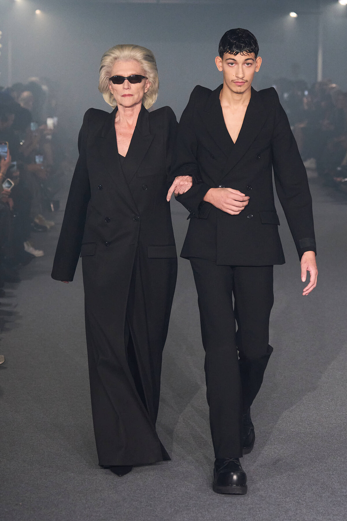 Two models walk down a runway arm-in-arm in matching black suits; an older woman with sunglasses on the left and a younger man on the right.