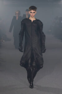 Model walks a futuristic black leather coat with padded shoulders down a smoky runway, dramatic makeup.