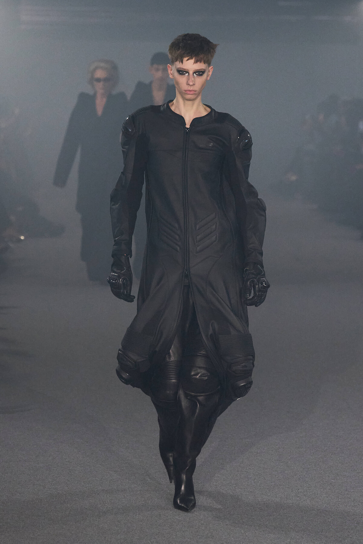 Model walks a futuristic black leather coat with padded shoulders down a smoky runway, dramatic makeup.