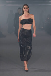 Runway model walking in a black leather strapless bandeau top and matching high-waisted leather skirt with sunglasses.