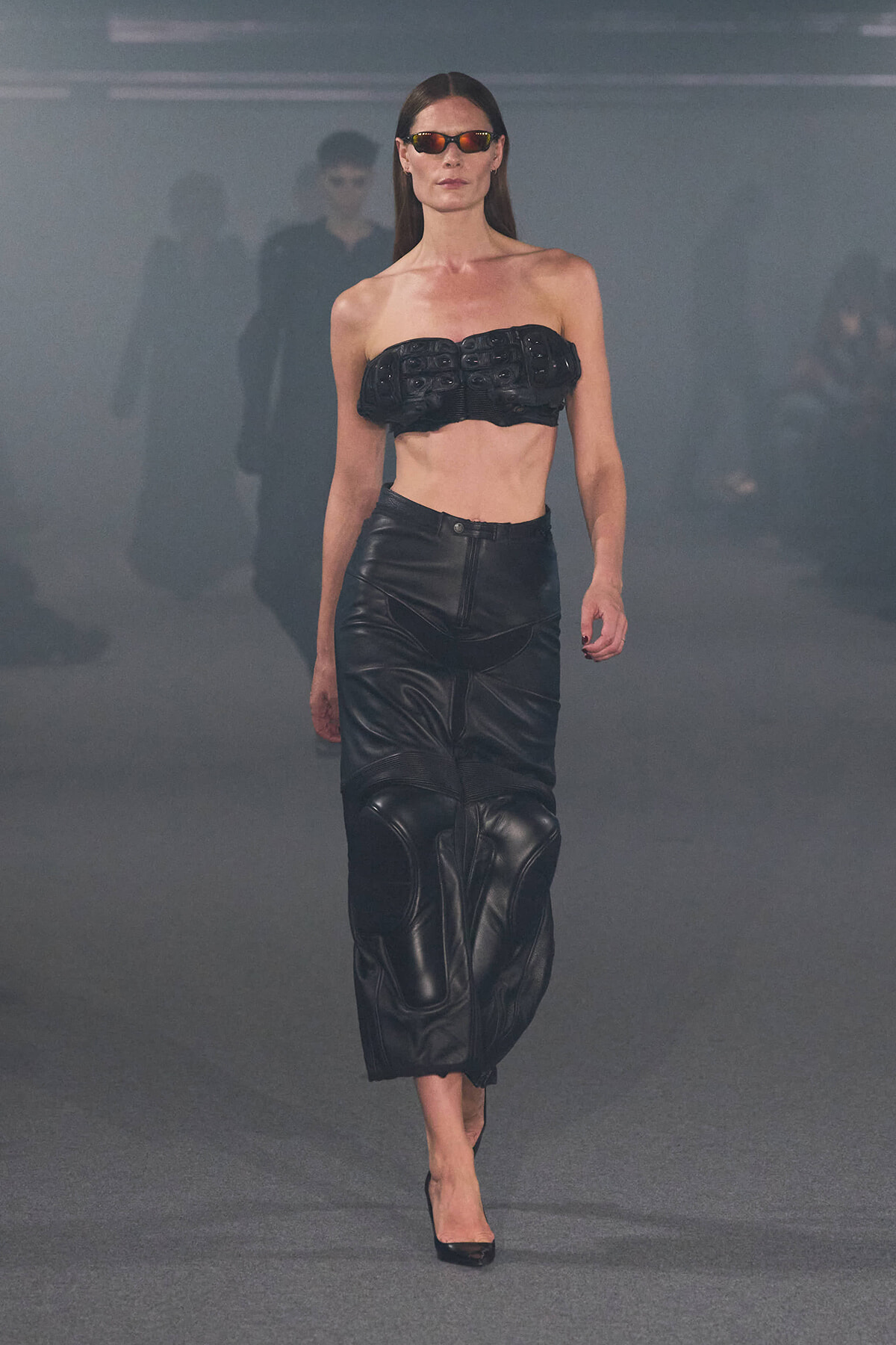 Runway model walking in a black leather strapless bandeau top and matching high-waisted leather skirt with sunglasses.