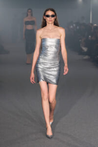 Model walking a runway in a strapless metallic silver mini dress and pointed heels, wearing dark sunglasses.