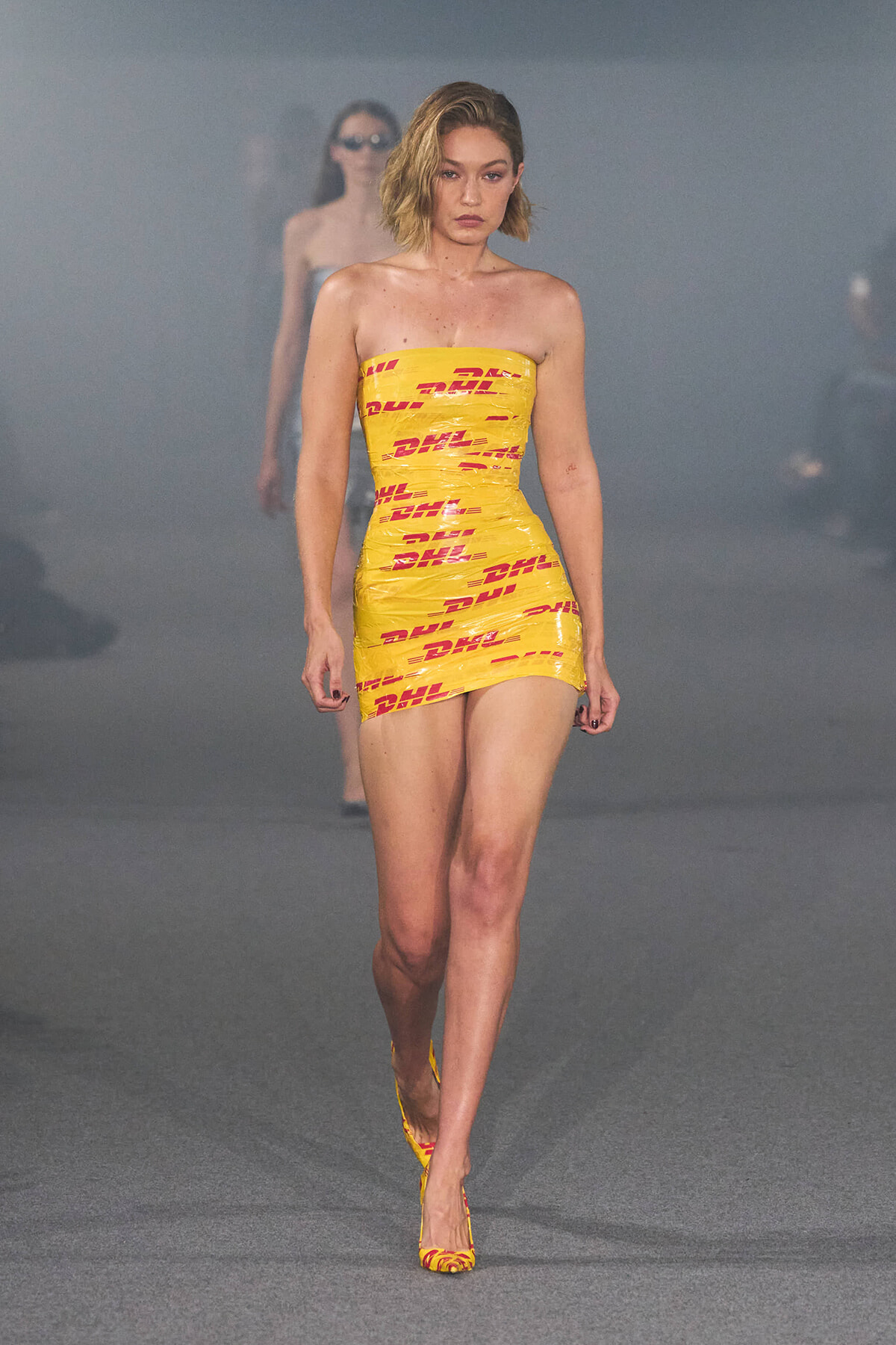 Model walks the runway in a yellow DHL-branded strapless mini dress and matching heels, gray foggy backdrop behind.