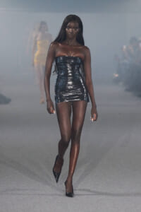 Model walks the runway in a strapless, shiny black mini dress with pointed heels amid a smoky backdrop.