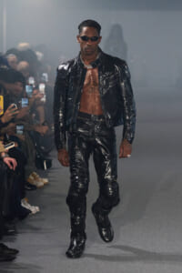 Male model in a glossy black patent leather jacket and pants, shirtless chest, wearing sunglasses and a silver chain on a runway.