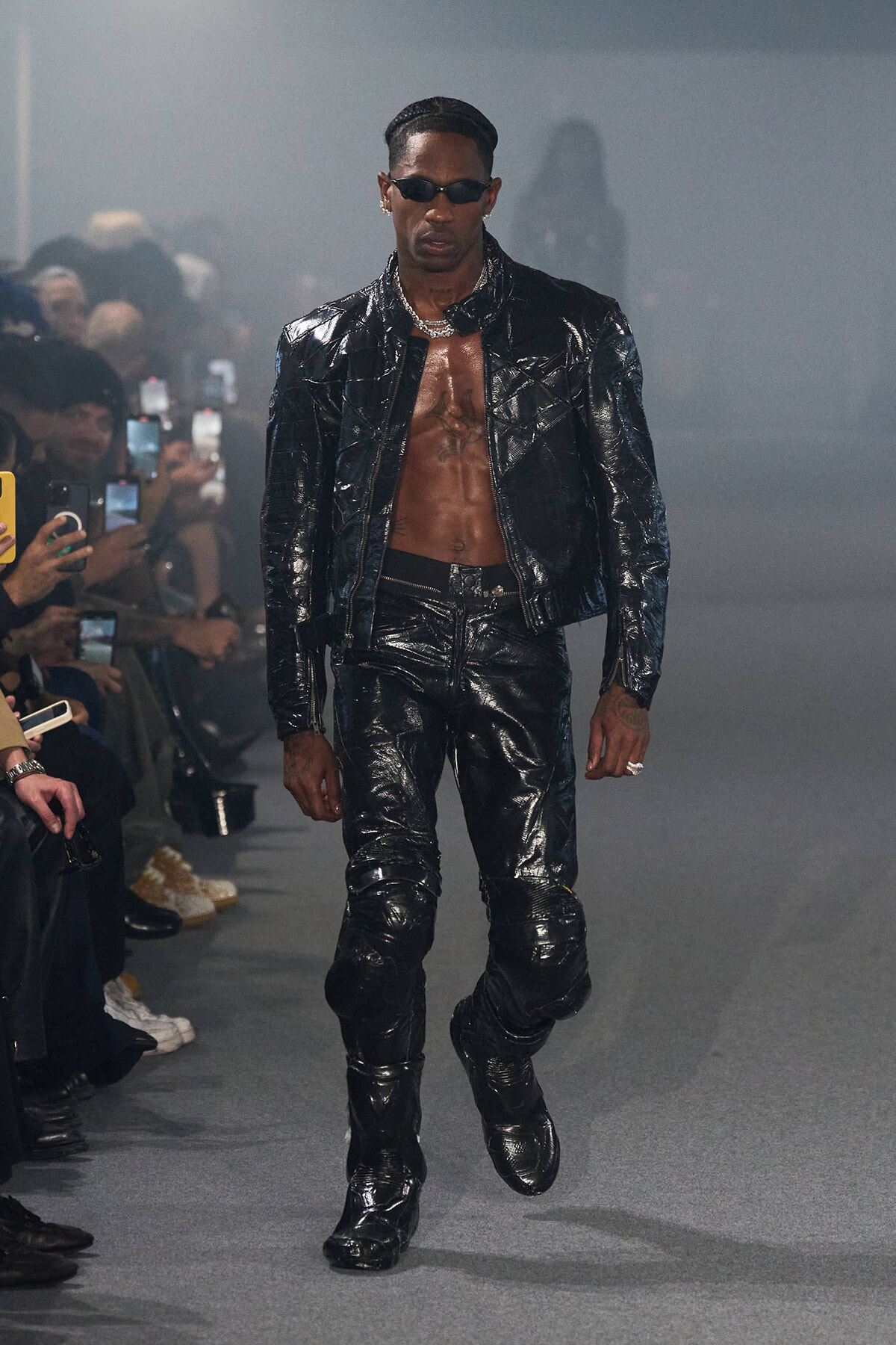 Male model in a glossy black patent leather jacket and pants, shirtless chest, wearing sunglasses and a silver chain on a runway.