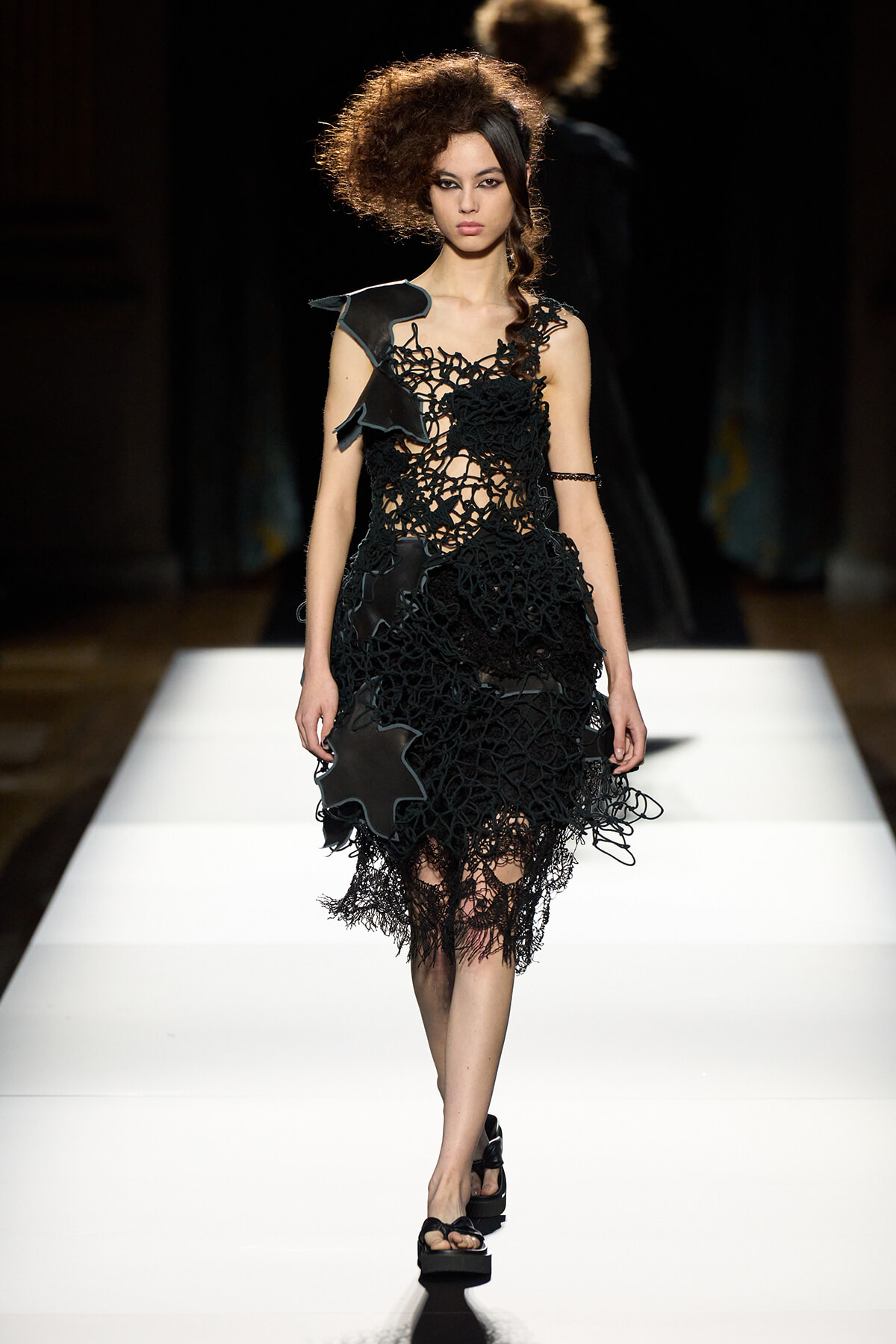 Model walks a black crochet dress with irregular floral motifs on a white runway. Voguish, avant-garde design with sculptural edges.