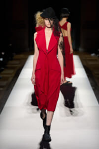 Model on the runway wearing a bold red sleeveless dress with a deep V, black hat, and a lace veil draped over one side of her face.