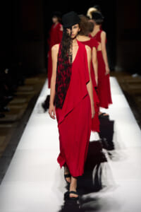 Runway model in a red draped dress with a black lace panel and a tilted black hat walking on a white catwalk.