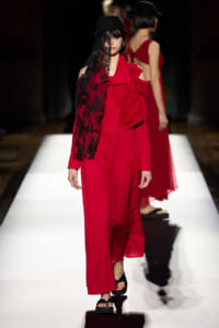 Model on a fashion runway wearing a red wide-leg jumpsuit with a black lace cape and a black hat.