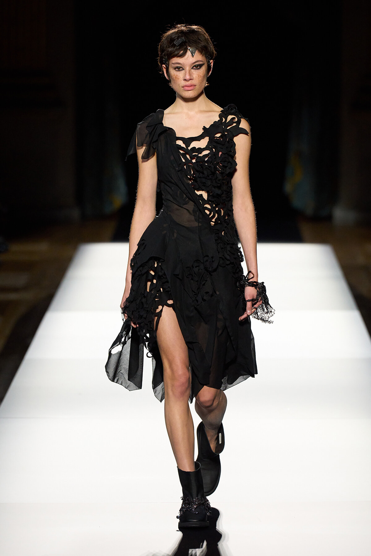 Model walking a runway in a black, intricately laced dress with asymmetric cutouts and sheer panels, on a white catwalk.