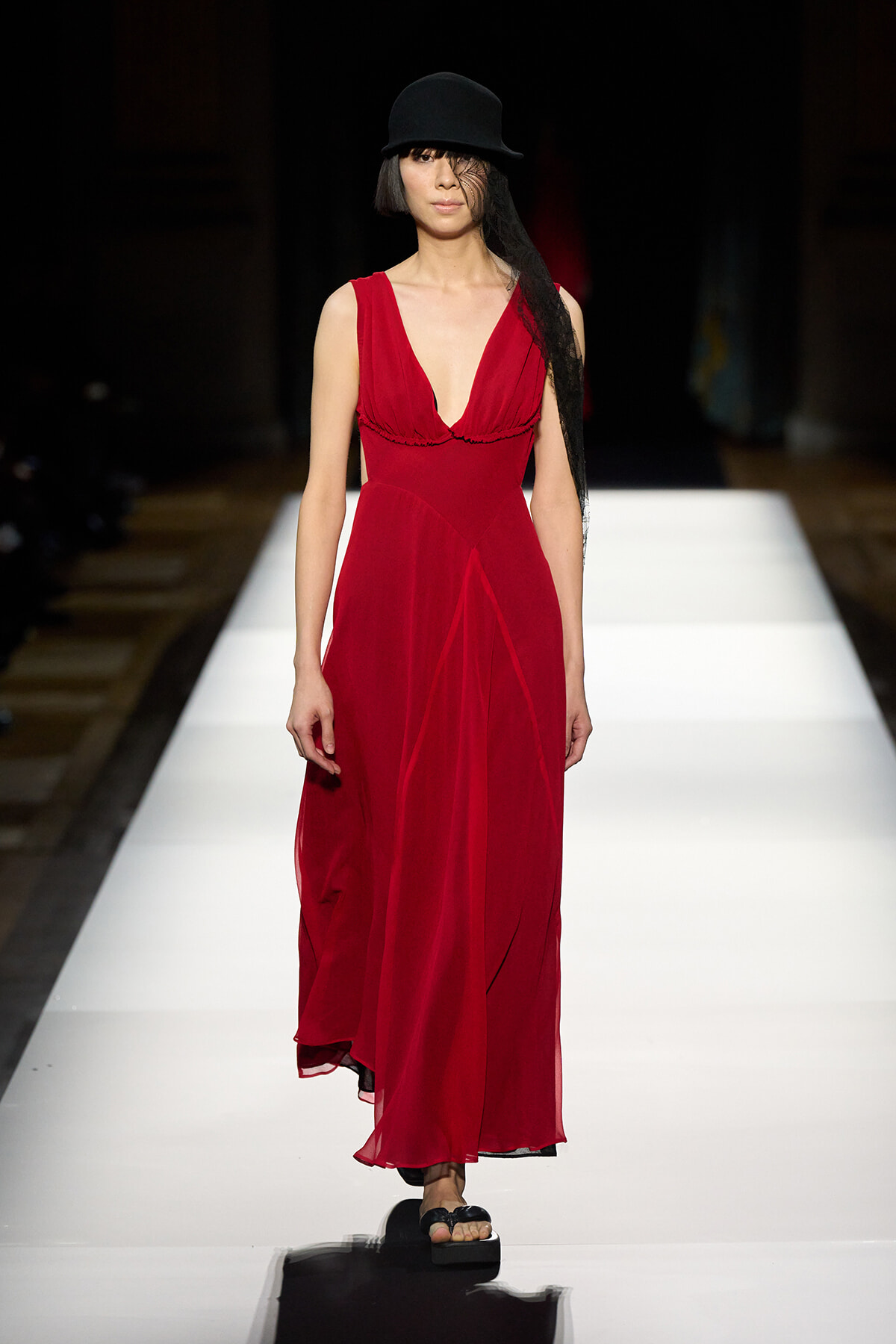 Model walks the runway in a long red V-neck chiffon dress with a black hat and veil accessory.