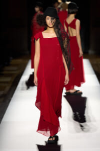 Model walking the runway in a red ankle-length dress with an asymmetric square neckline, black hat, and lace veil draped over her face