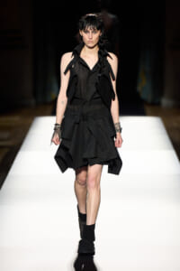 Model walking a fashion runway in a black layered sleeveless dress with a wide collar and strap details, paired with black ankle boots.