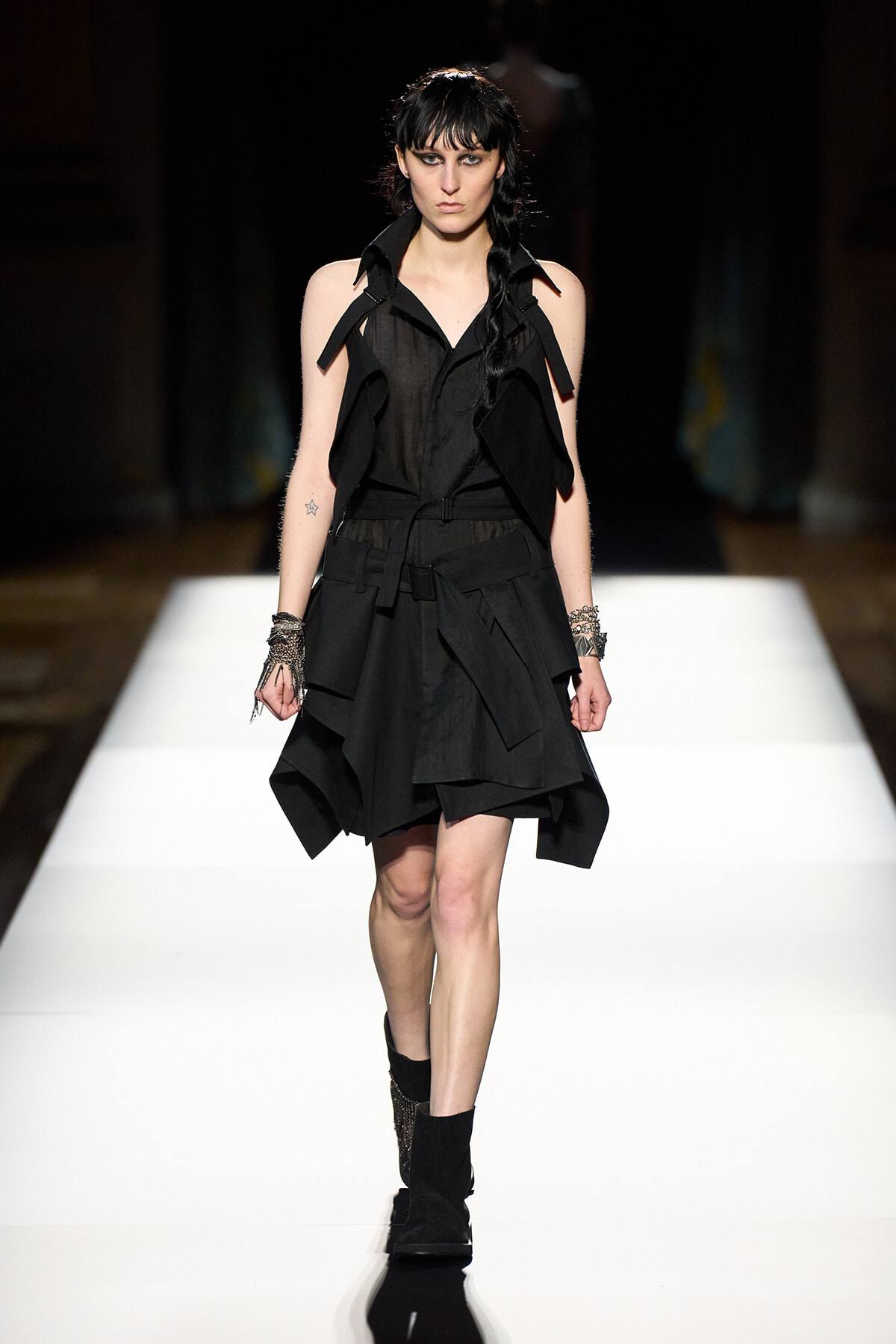 Model walking a fashion runway in a black layered sleeveless dress with a wide collar and strap details, paired with black ankle boots.