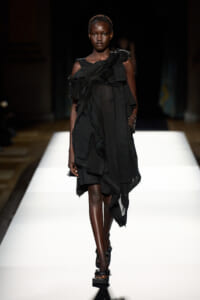 Black-skinned model walking a runway in a layered, black draped dress with an asymmetric hem and ruffles.
