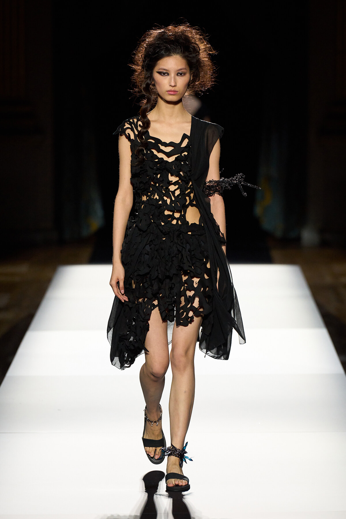 Runway model in a black, sculptural cut‑out dress with layered, irregular shapes walking a white catwalk.