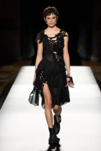 Model walks a runway in a black gothic dress with floral cutout detailing, sheer panels, and high-low hem, paired with black ankle boots.