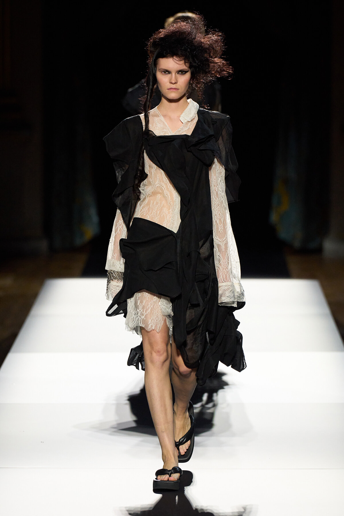 Model walking a runway in a black and white lace avant-garde dress with layered draped fabric and sandals on a white catwalk.