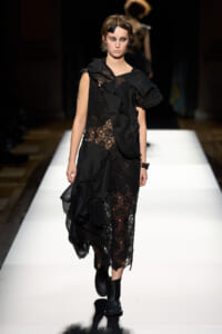 Model walks the runway in a black lace dress with a ruffled overlay and ankle boots, under dramatic lighting.