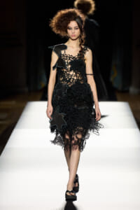 Runway model in a black, crochet-inspired dress with lace overlays walking a white catwalk; dramatic voluminous hairstyle adds height to the look, fashion show scene.