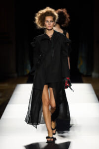 Model walking the runway in a black layered coat with a sheer overlay, black sandals, and a voluminous, messy hairstyle.