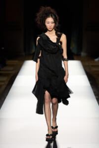 Female model walking a bright white runway in a black, layered, draped dress with ruffled sleeves and open-toe sandals.