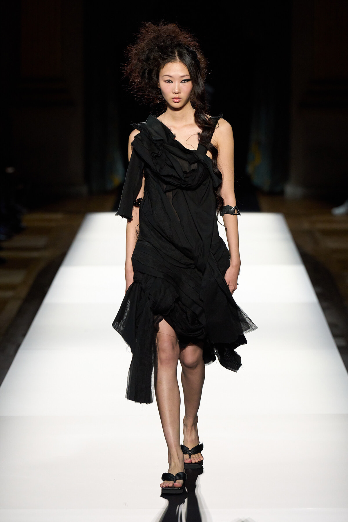 Female model walking a bright white runway in a black, layered, draped dress with ruffled sleeves and open-toe sandals.