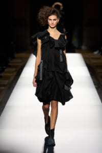 Model walking a white runway in a black, ruffled dress with gray ribbon accents and black ankle boots.